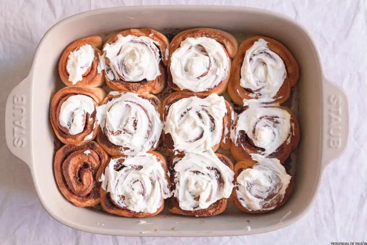 Horneados rollitos de canela cubiertos con glaseado cremoso en un molde de cerámica.