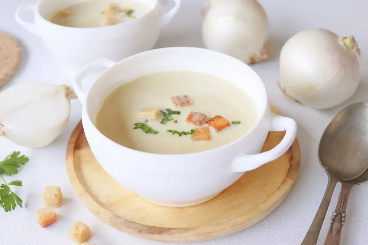 Crema de cebolla cremosa, adornada con picatostes dorados y perejil fresco. Cebollas blancas enteras y cortadas a medias rodean los cuencos.