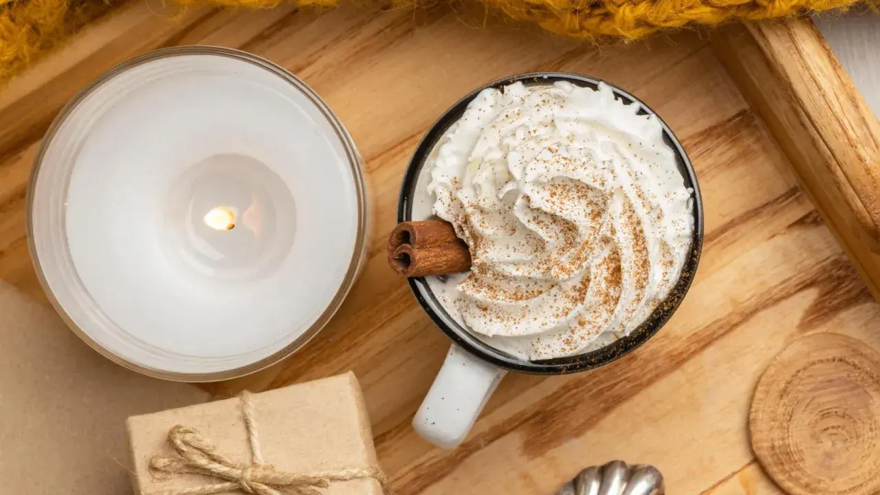 Taza de bebida caliente con abundante **montar nata**, canela y una rama de canela. Una vela encendida y un regalo completan la escena acogedora.