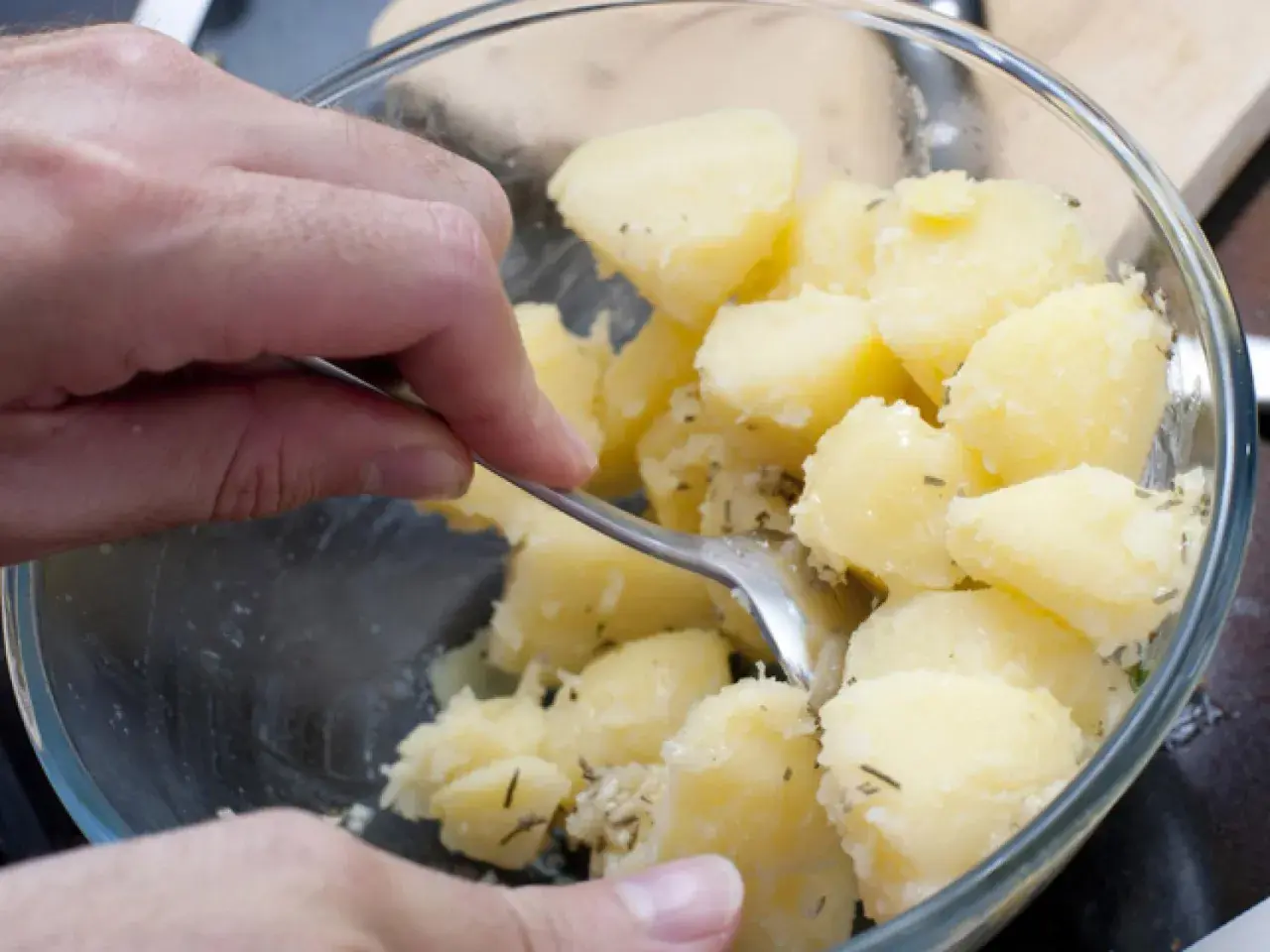 Manos mezclando patatas cocidas con hierbas en un bol de cristal.