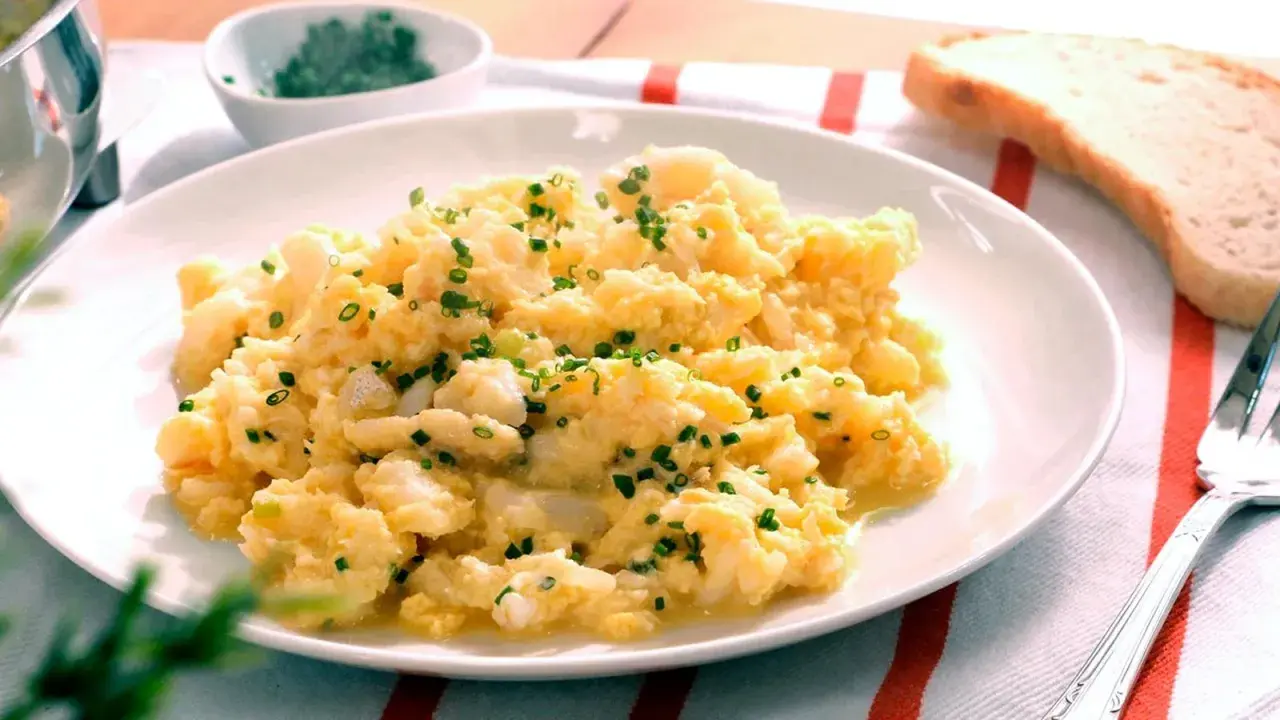 Revuelto de bacalao cremoso, espolvoreado con cebollino fresco, servido en un plato blanco con una rebanada de pan tostado al lado.