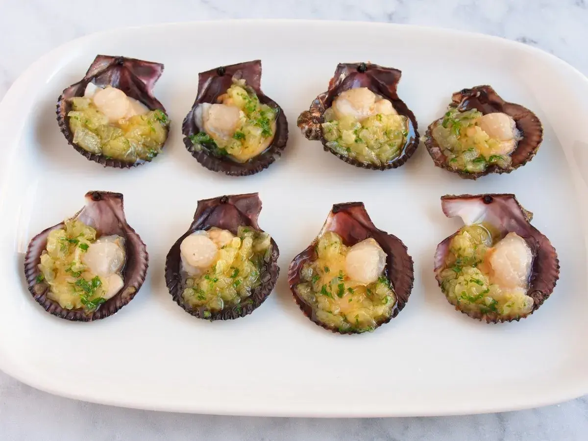 Ocho zamburiñas al horno, listas para disfrutar, con un relleno dorado y perejil picado.