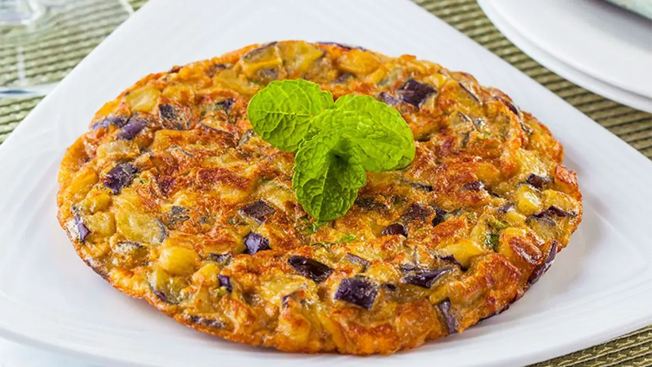 Tortilla de berenjena dorada y esponjosa, con trozos morados de berenjena y patata, decorada con hojas verdes de menta.