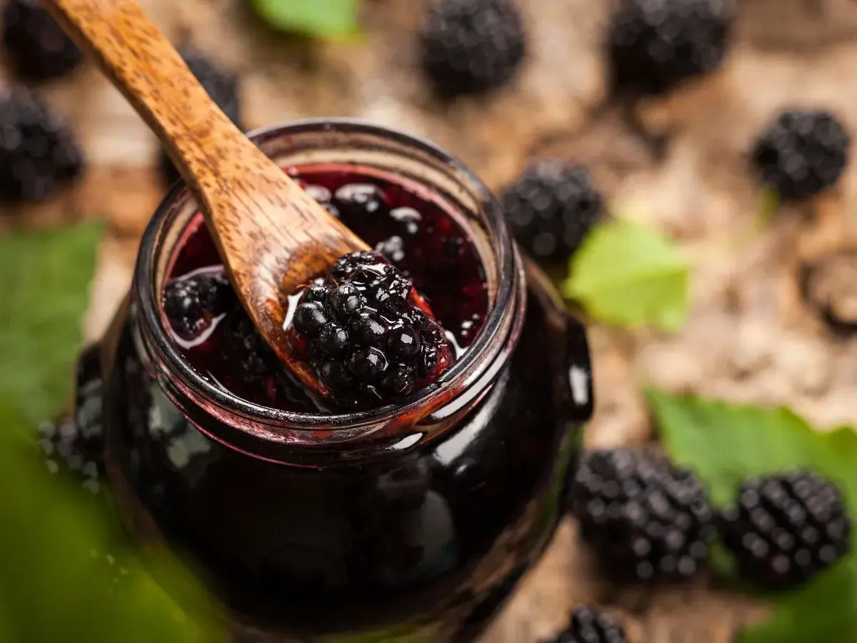 Cuchara de madera recoge mermelada de moras silvestres de un tarro, rodeado de moras frescas y hojas verdes.