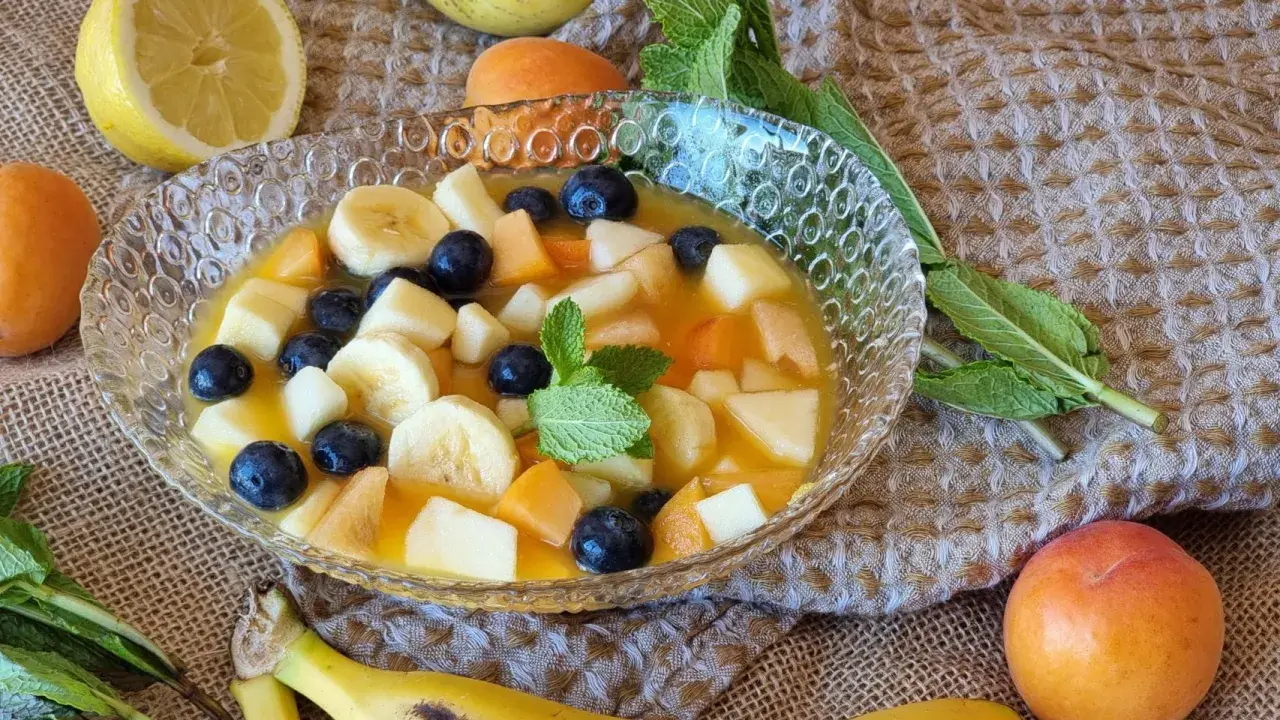 Deliciosa macedonia de frutas con plátano, arándanos, melocotón y manzana en un cuenco de cristal.