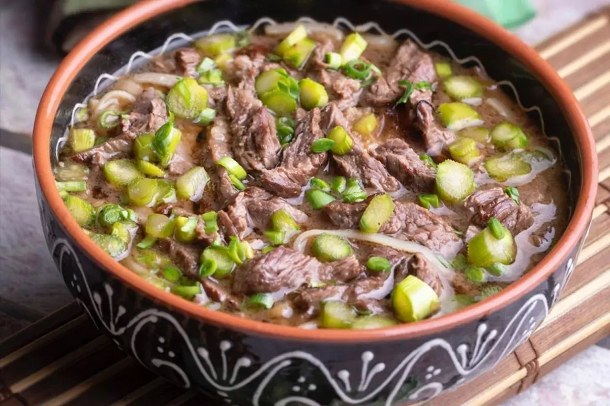 Un reconfortante plato de ternera guisada con trozos de carne tierna, fideos y espárragos verdes, servido en un cuenco rústico.