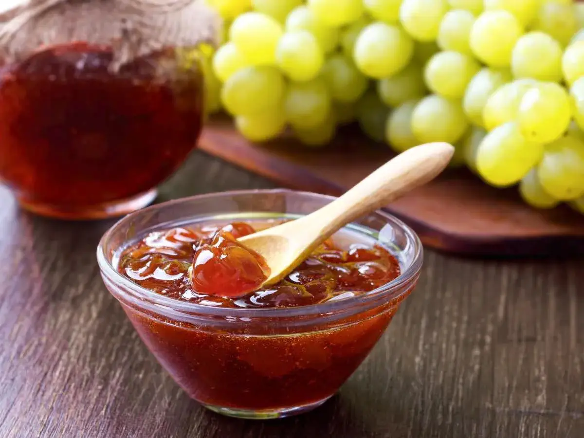 Un cuenco de cristal con mermelada de uvas casera y una cuchara de madera, junto a un racimo de uvas verdes.