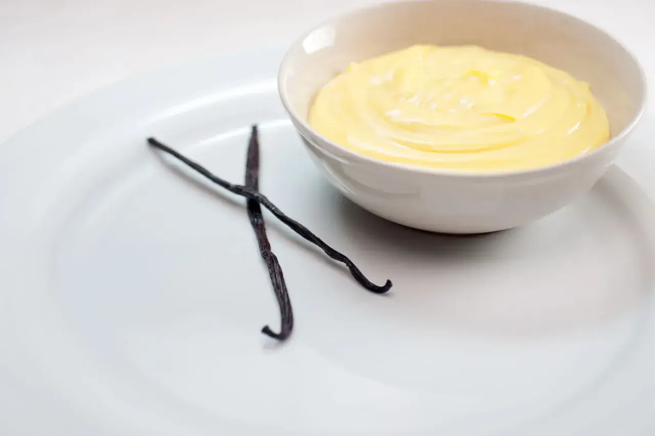 Crema inglesa cremosa en un tazón, con dos vainas de vainilla oscuras al lado.