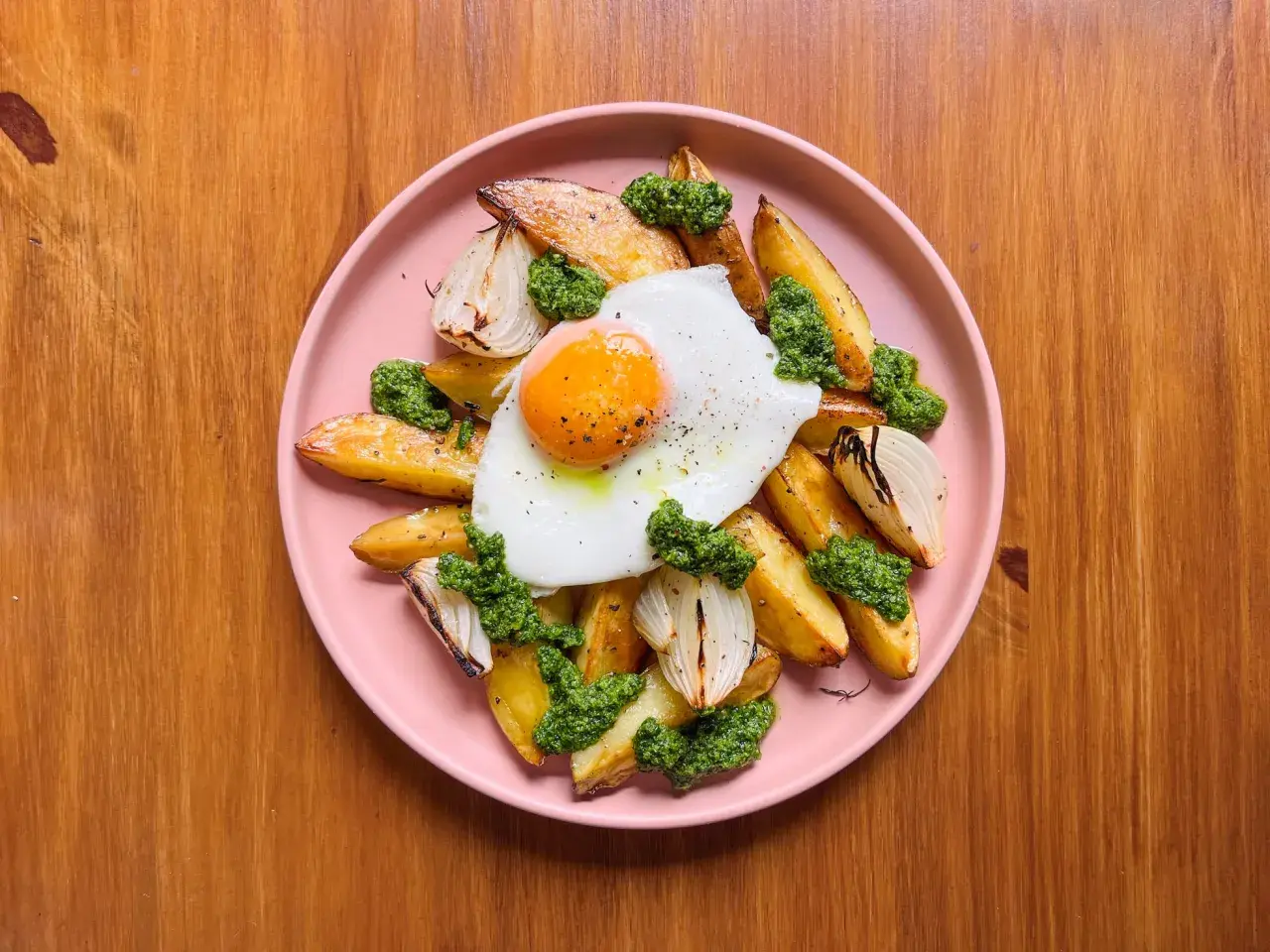 Patatas al horno doradas con huevo frito, cebolla asada y pesto verde, servidas en un plato rosa sobre una mesa de madera.