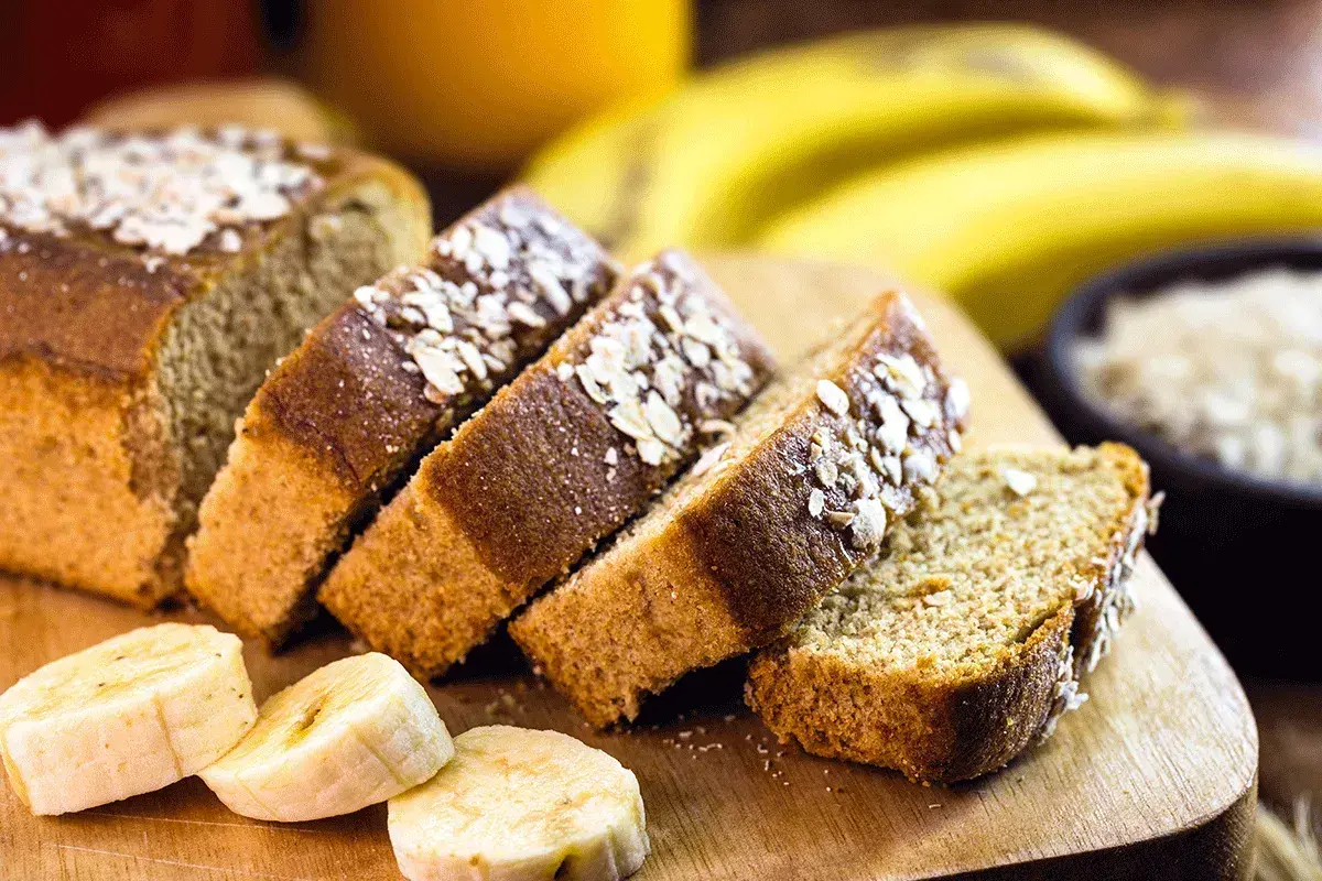 Rebanadas de bizcocho de plátano sin azúcar, cubiertas con avena, junto a plátanos frescos y un tazón de avena.