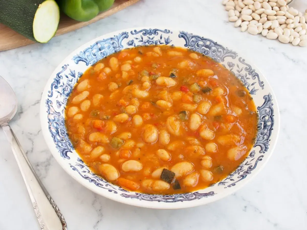 Un plato de alubias blancas con verduras, cocinadas en una salsa sabrosa. Se ven trozos de pimiento, zanahoria y calabacín.