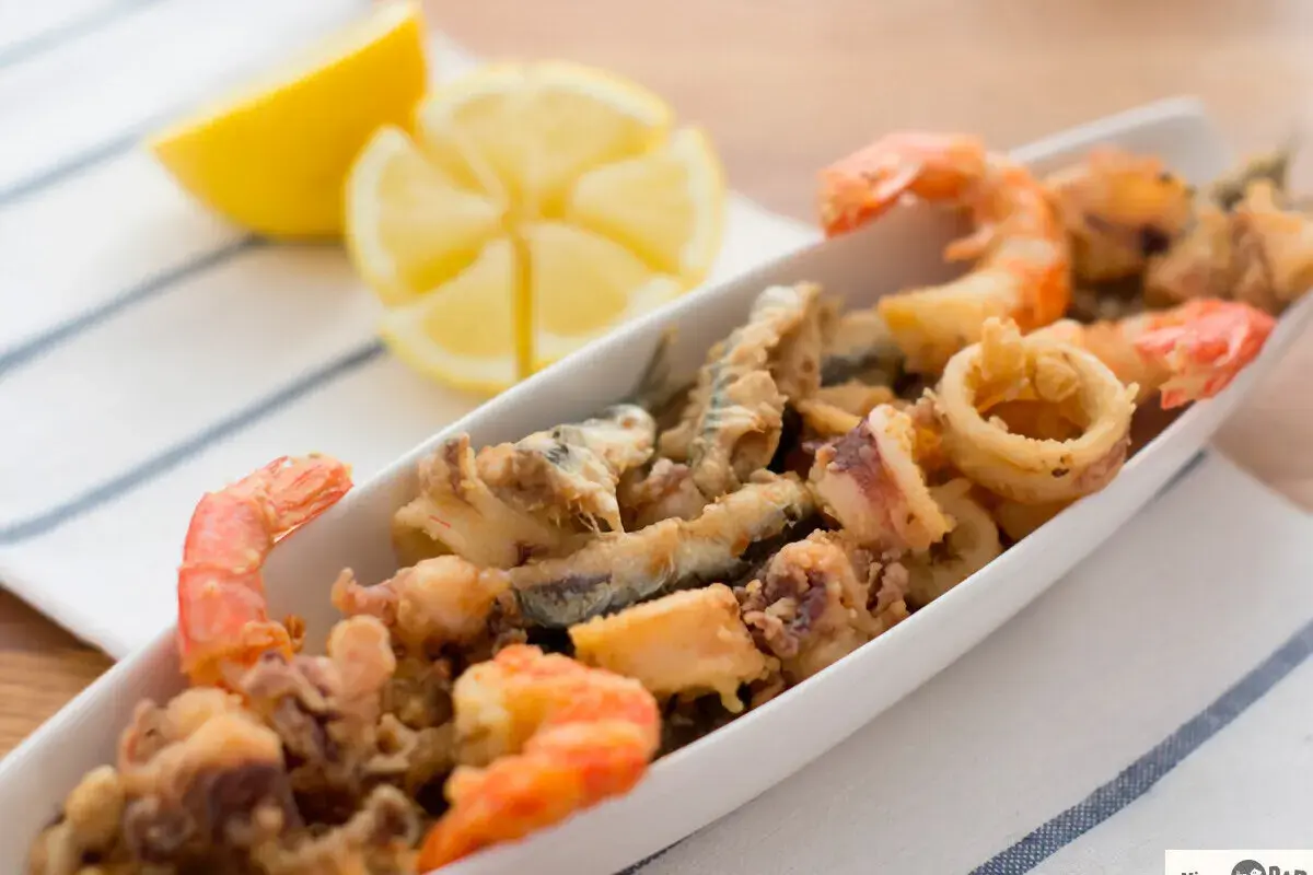 Deliciosa fritura de pescado con gambas y calamares, servida en una bandeja blanca con gajos de limón.