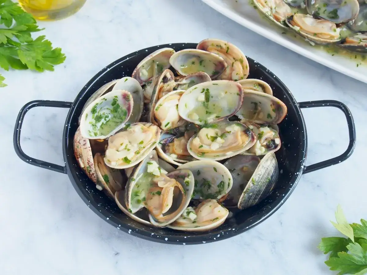 Almejas en salsa verde, con ajo picado y perejil fresco, servidas en una sartén negra.