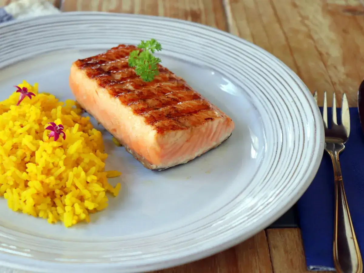 Un delicioso salm&oacute;n a la plancha con marcas de parrilla, servido con arroz amarillo y decorado con perejil y flores moradas.