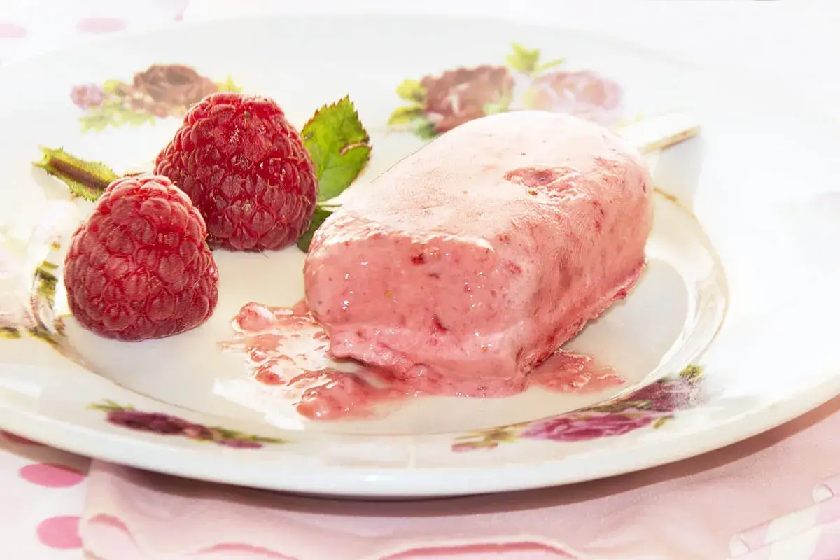 Helado de frambuesa con dos frambuesas frescas al lado. La dulzura perfecta para un día caluroso, hecha con azúcar invertida.