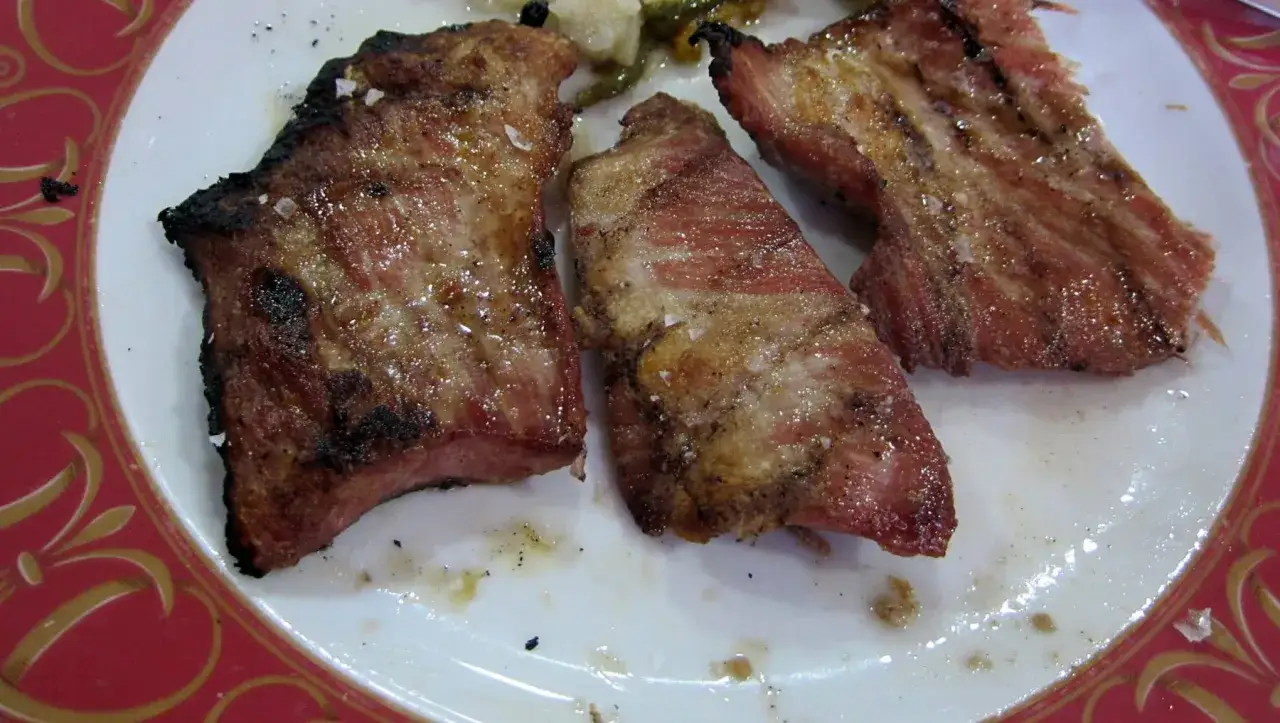 Tres trozos de churrasco de cerdo a la parrilla, jugosos y con sal gruesa, servidos en un plato blanco con borde rojo y dorado.