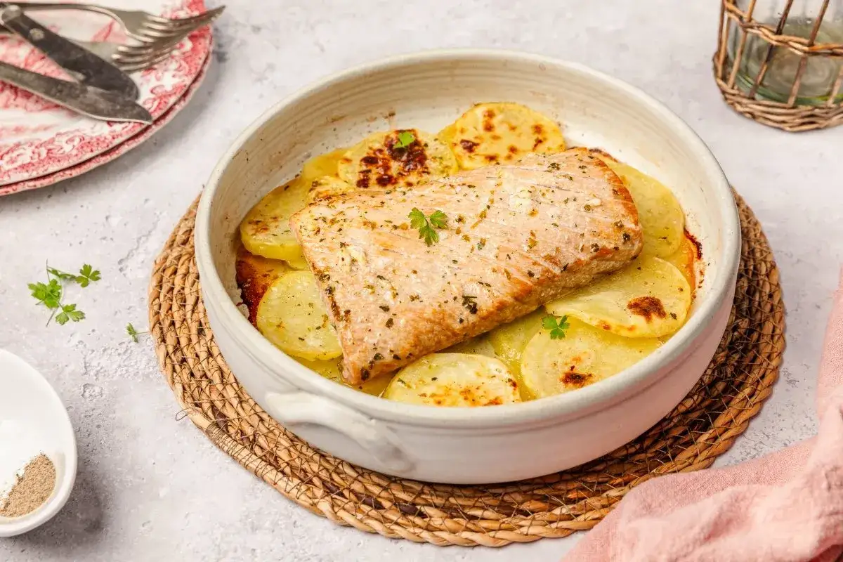 Deliciosa ventresca al horno con patatas laminadas y hierbas aromáticas, servida en una cazuela de barro sobre una esterilla de mimbre.
