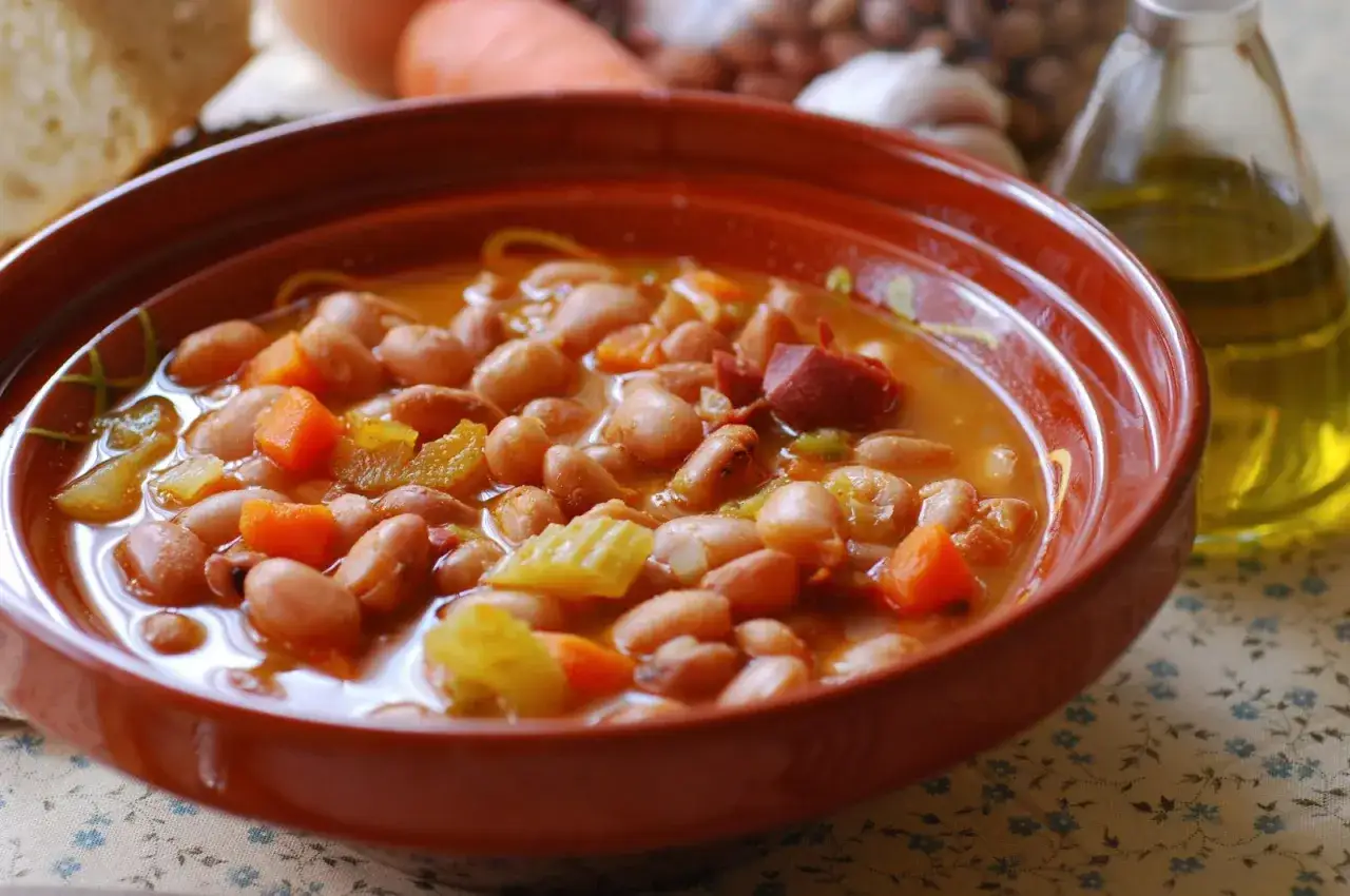 Un plato humeante de alubias pintas con trozos de chorizo y verduras, acompañado de aceite de oliva.