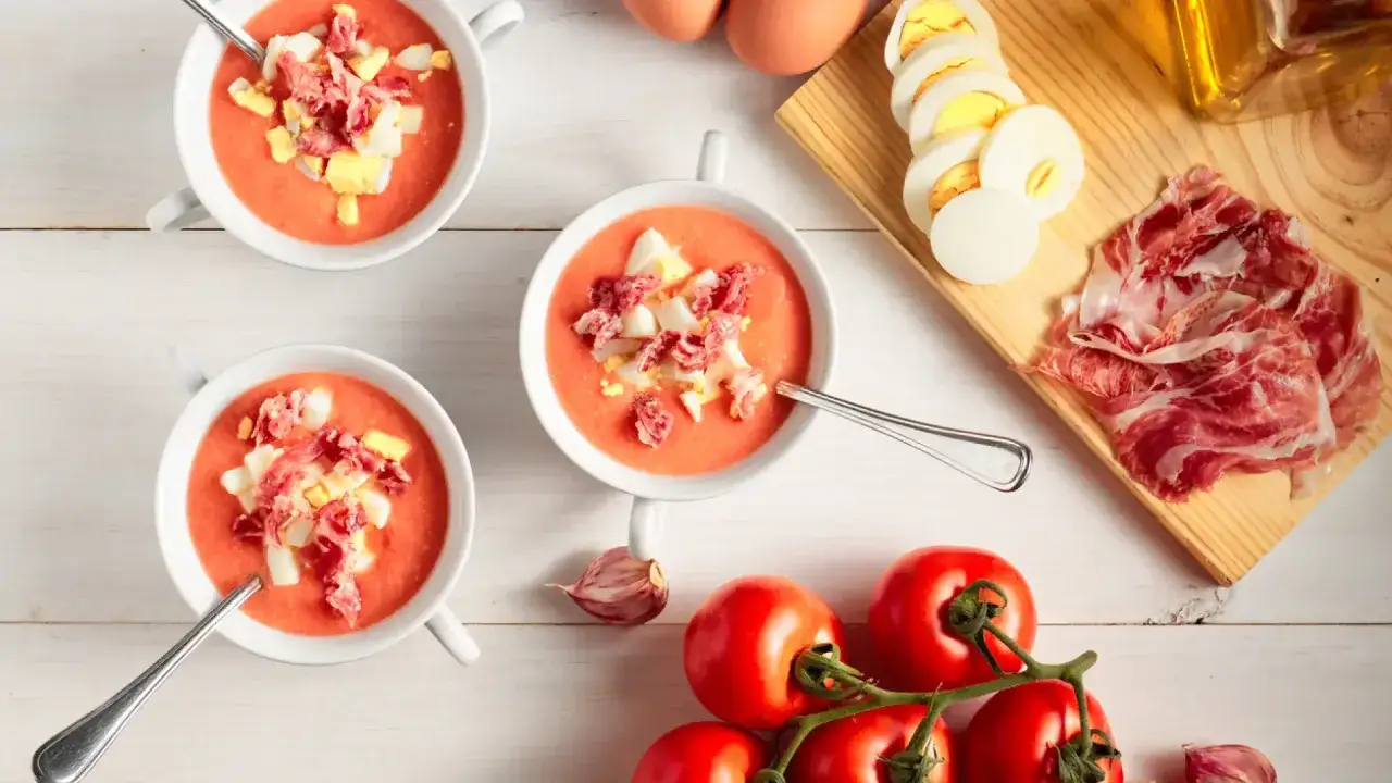 Salmorejo cremoso hecho en Thermomix, servido en cuencos blancos con huevo duro y jam&oacute;n picado. Tomates frescos y ajo en la mesa.