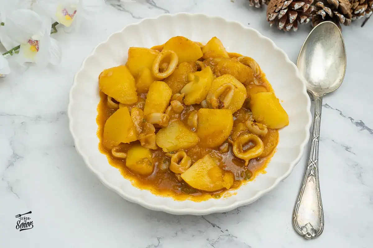 Plato de patatas con calamares en salsa, servido en un plato blanco con una cuchara antigua.