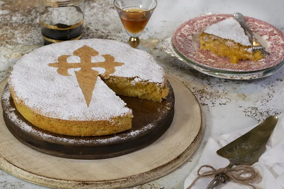 Tarta de almendras espolvoreada con az&uacute;car glas, con la Cruz de Santiago grabada. Una porci&oacute;n ya servida en un plato.