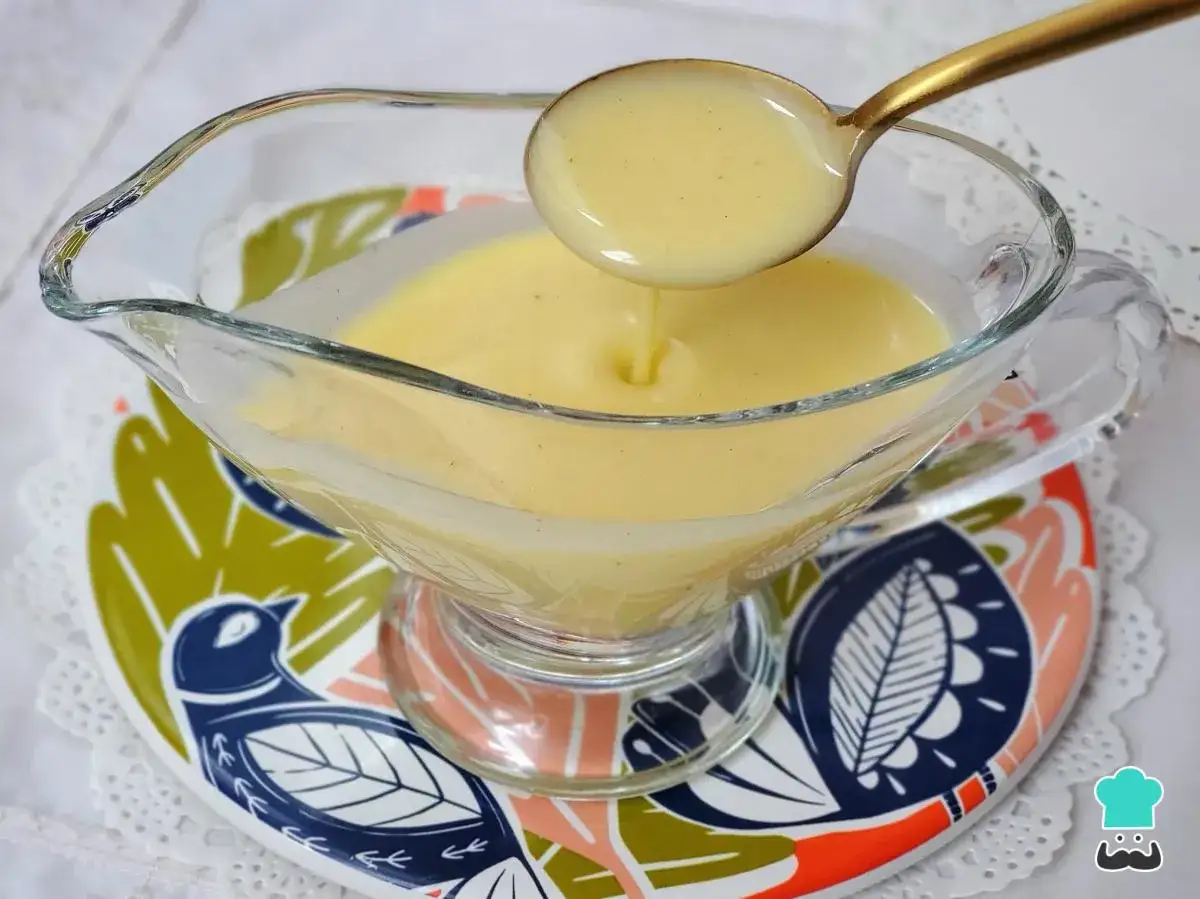Cucharada de cremosa crema inglesa cayendo en una salsera de cristal, sobre un plato decorado con pájaros y hojas.
