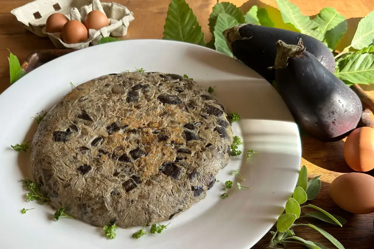 Tortilla de berenjena r&uacute;stica, con trozos visibles de la verdura, acompa&ntilde;ada de huevos frescos y hojas verdes.