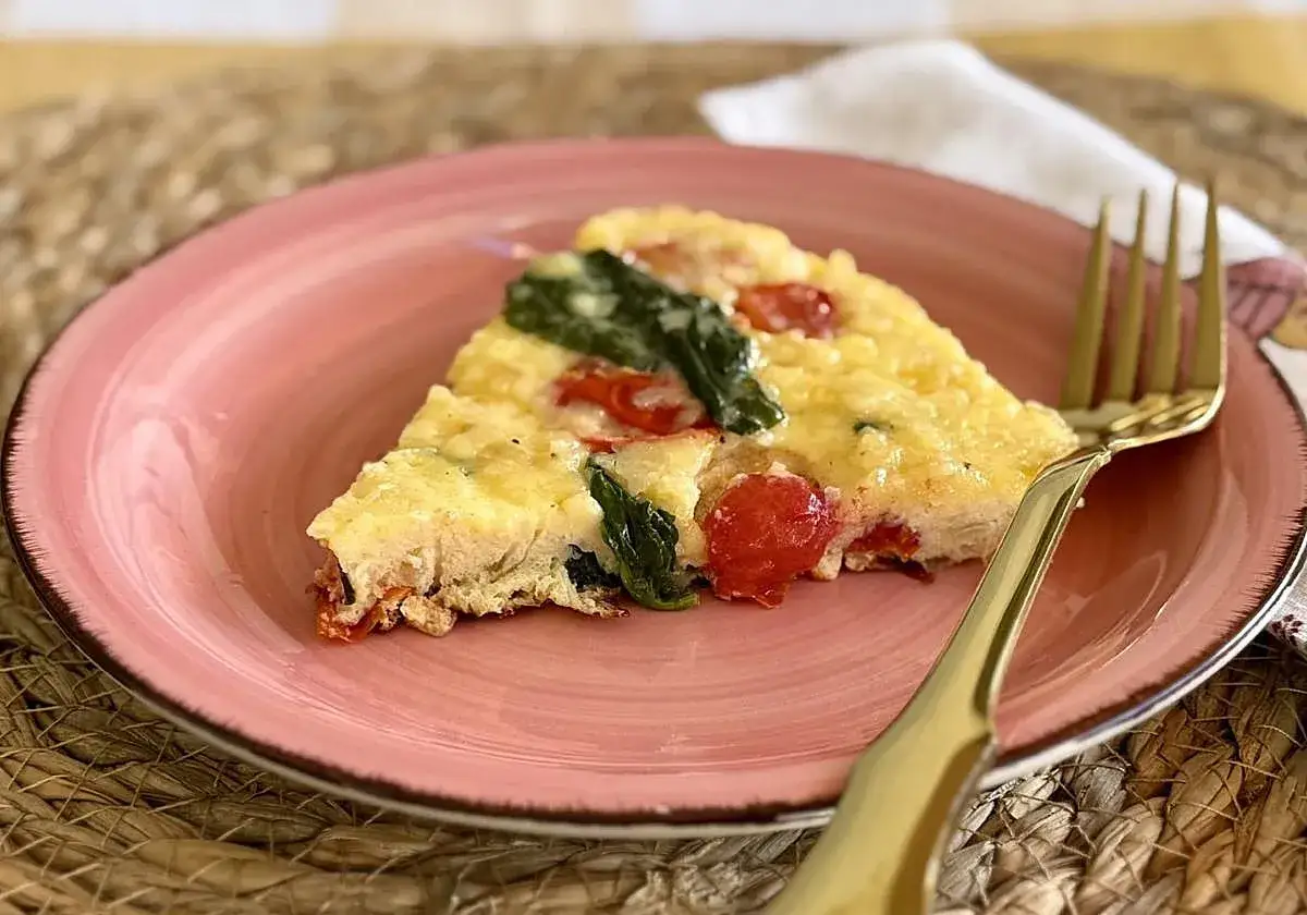 Rebanada de frittata de verduras con tomates cherry y espinacas, servida en plato rosa con tenedor dorado.