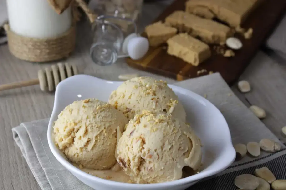Delicioso helado de turr&oacute;n con trocitos de almendra, servido en un cuenco blanco. Al fondo, barras de turr&oacute;n y almendras.