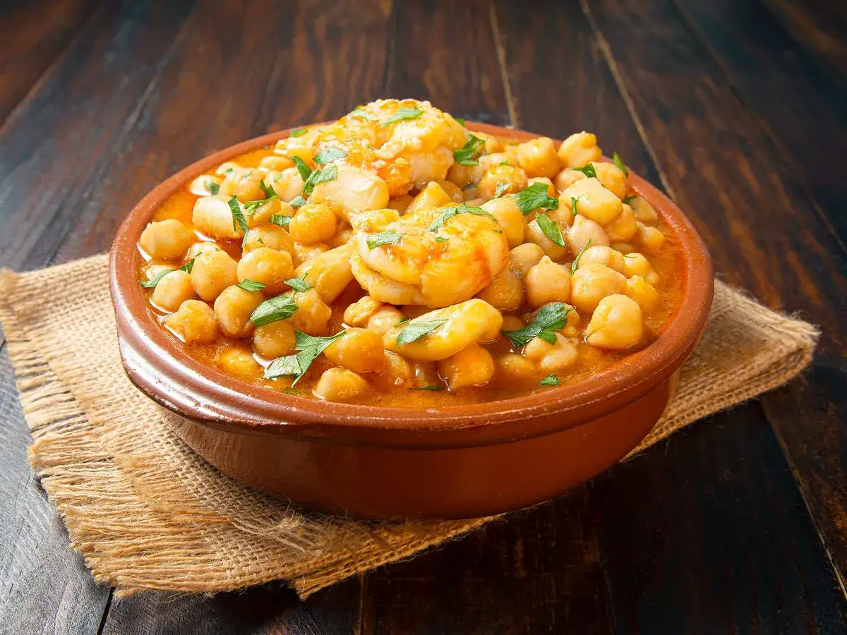 Un tazón de barro con garbanzos con langostinos, adornado con perejil fresco, sobre una tela de arpillera y una mesa de madera oscura.