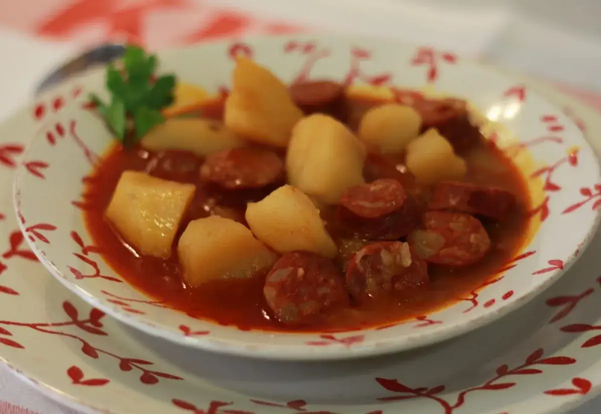 Un plato humeante de patatas a la riojana, con trozos de chorizo y patata en una salsa rojiza, decorado con una ramita de perejil.