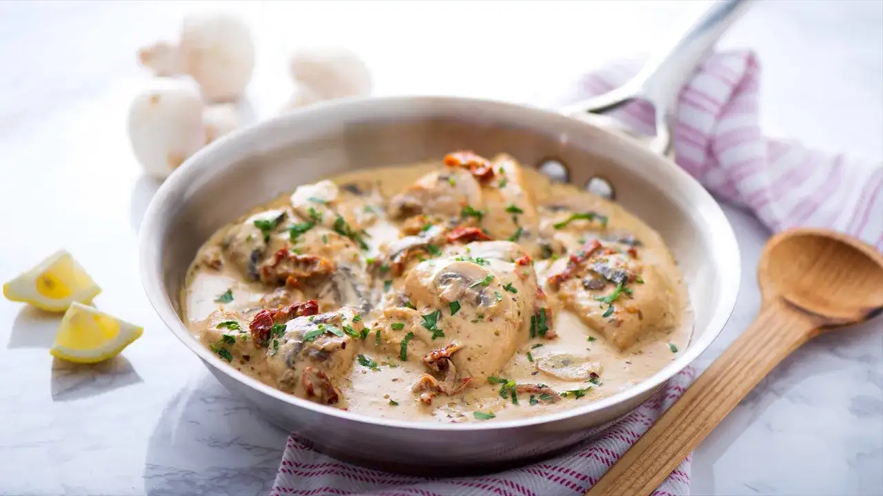Delicioso pollo con champi&ntilde;ones en salsa cremosa, adornado con perejil fresco y tomates secos. Servido en sart&eacute;n con rodajas de lim&oacute;n y cuchara de madera.