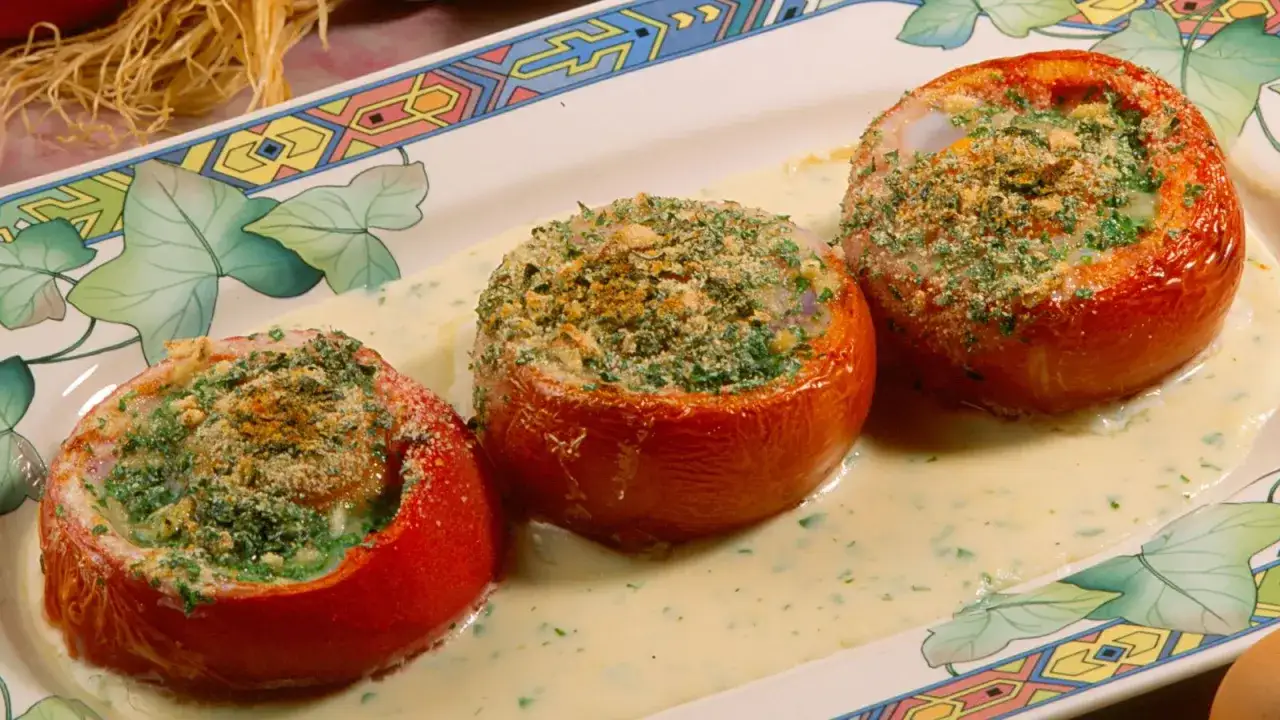 Tres tomates al horno rellenos con huevo y hierbas, servidos en una cremosa salsa sobre un plato decorado.