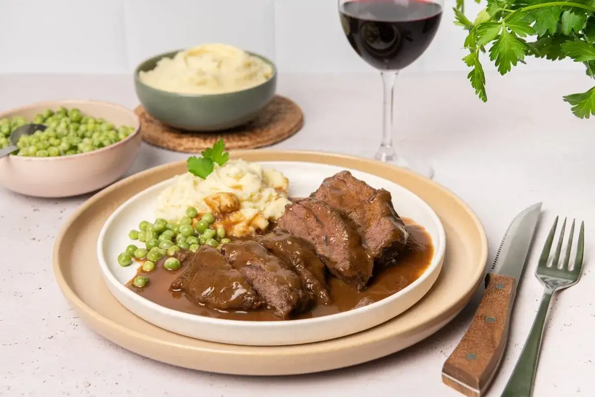 Carrilleras de ternera tiernas en salsa, servidas con puré de patatas y guisantes. Un plato reconfortante acompañado de vino tinto.