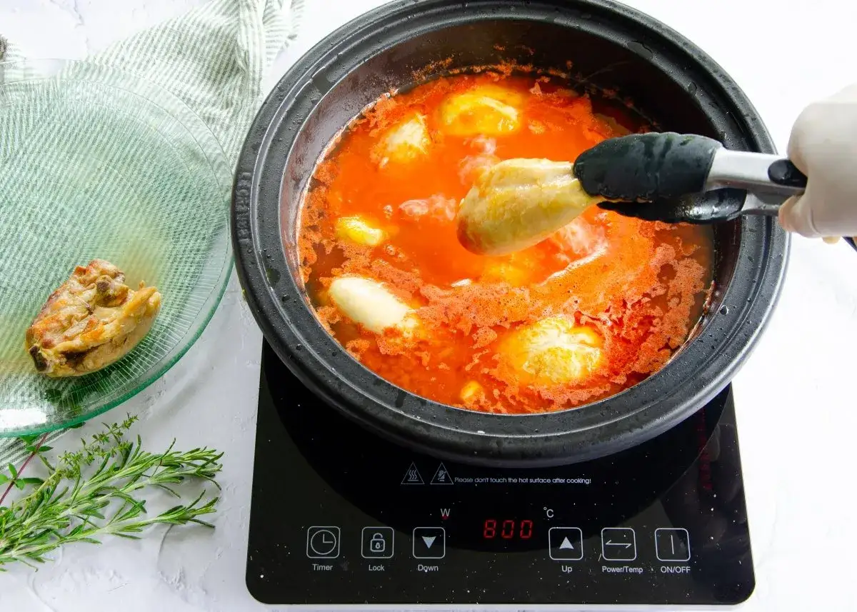 Un trozo de pollo se añade a una olla con salsa de tomate y otras piezas de pollo.