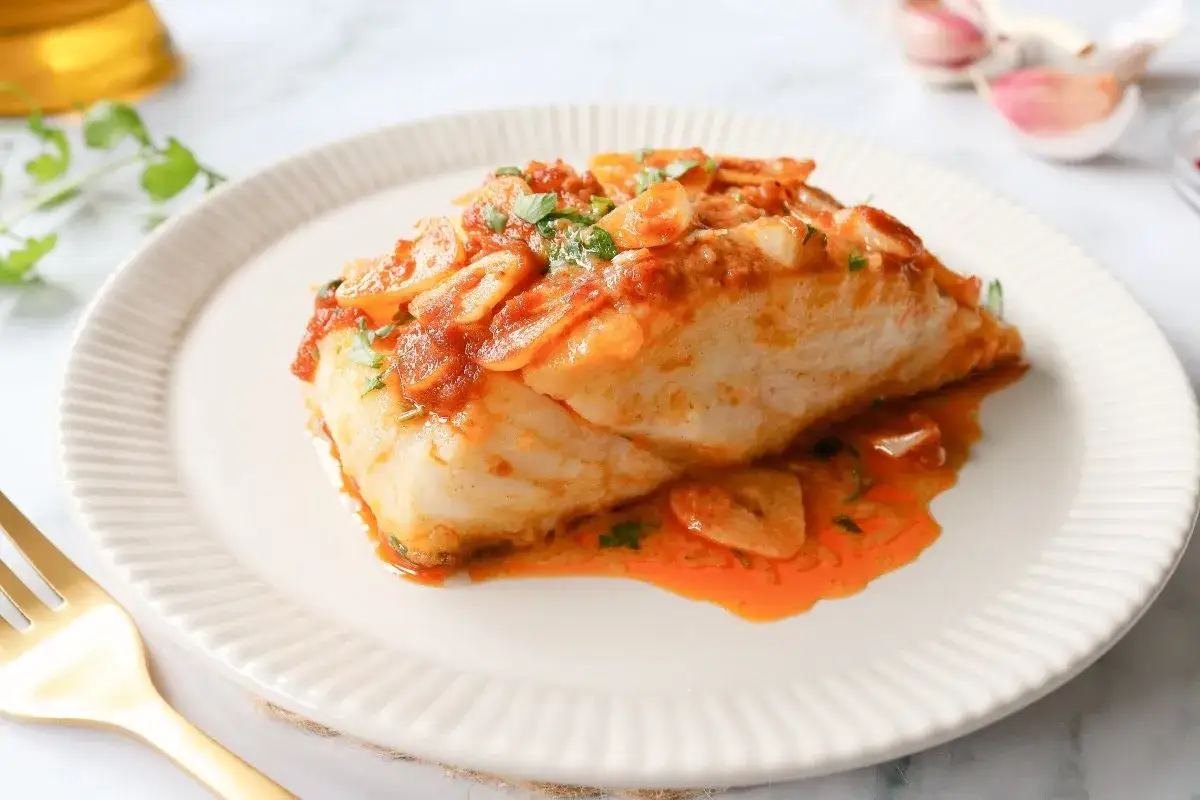 Bacalao a la llauna jugoso, cubierto de salsa de tomate y ajo laminado, servido en plato blanco con tenedor dorado.
