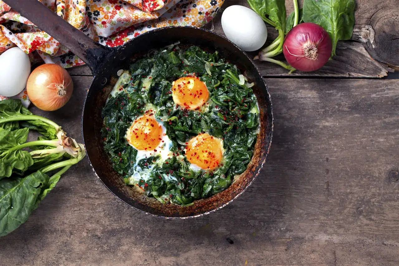 Espinacas salteadas con huevos rotos en sartén rústica, acompañadas de cebolla y espinacas frescas.