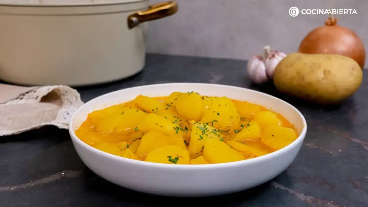 Un taz&oacute;n blanco lleno de deliciosas patatas guisadas, adornadas con perejil fresco. Al fondo, ingredientes para la receta: patatas, ajo y cebolla.