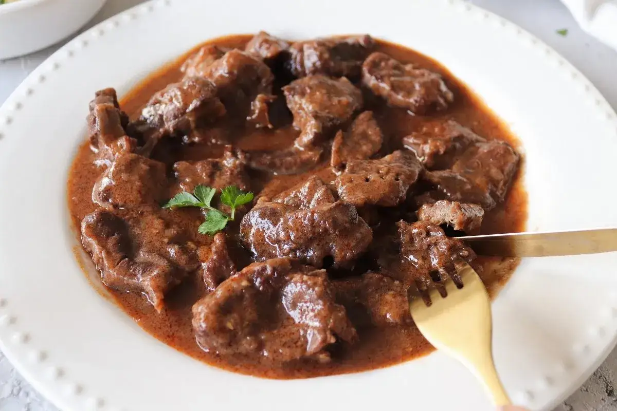 Plato de morcillo de ternera estofado en salsa oscura, servido en una fuente blanca con perejil. Un tenedor dorado corta un trozo.