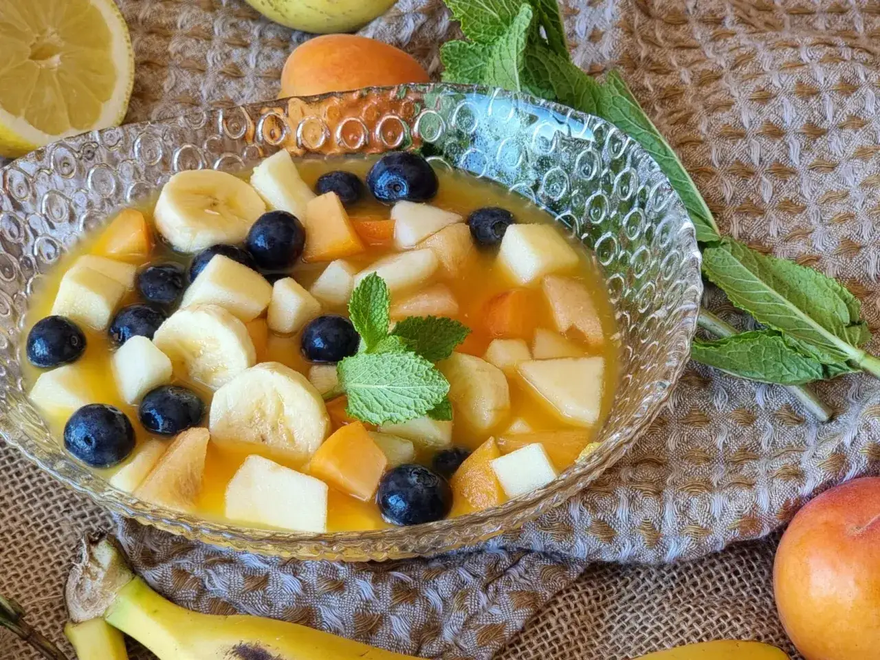 Deliciosa macedonia de frutas con pl&aacute;tano, ar&aacute;ndanos, melocot&oacute;n y manzana en un cuenco de cristal, decorada con menta.