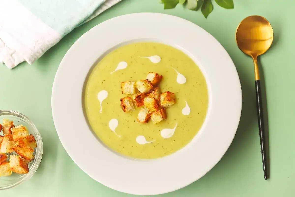 Crema de calabacín servida en plato blanco, decorada con picatostes y un chorrito de nata. Una cuchara dorada y un cuenco con más picatostes completan la escena.