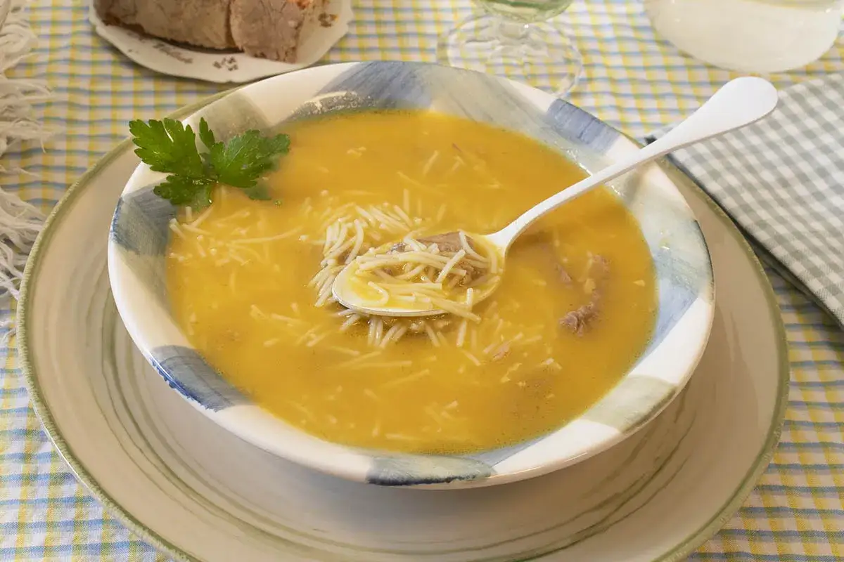 Un plato humeante de sopa de fideos con trozos de carne y perejil fresco. Una cuchara recoge un bocado sustancioso.