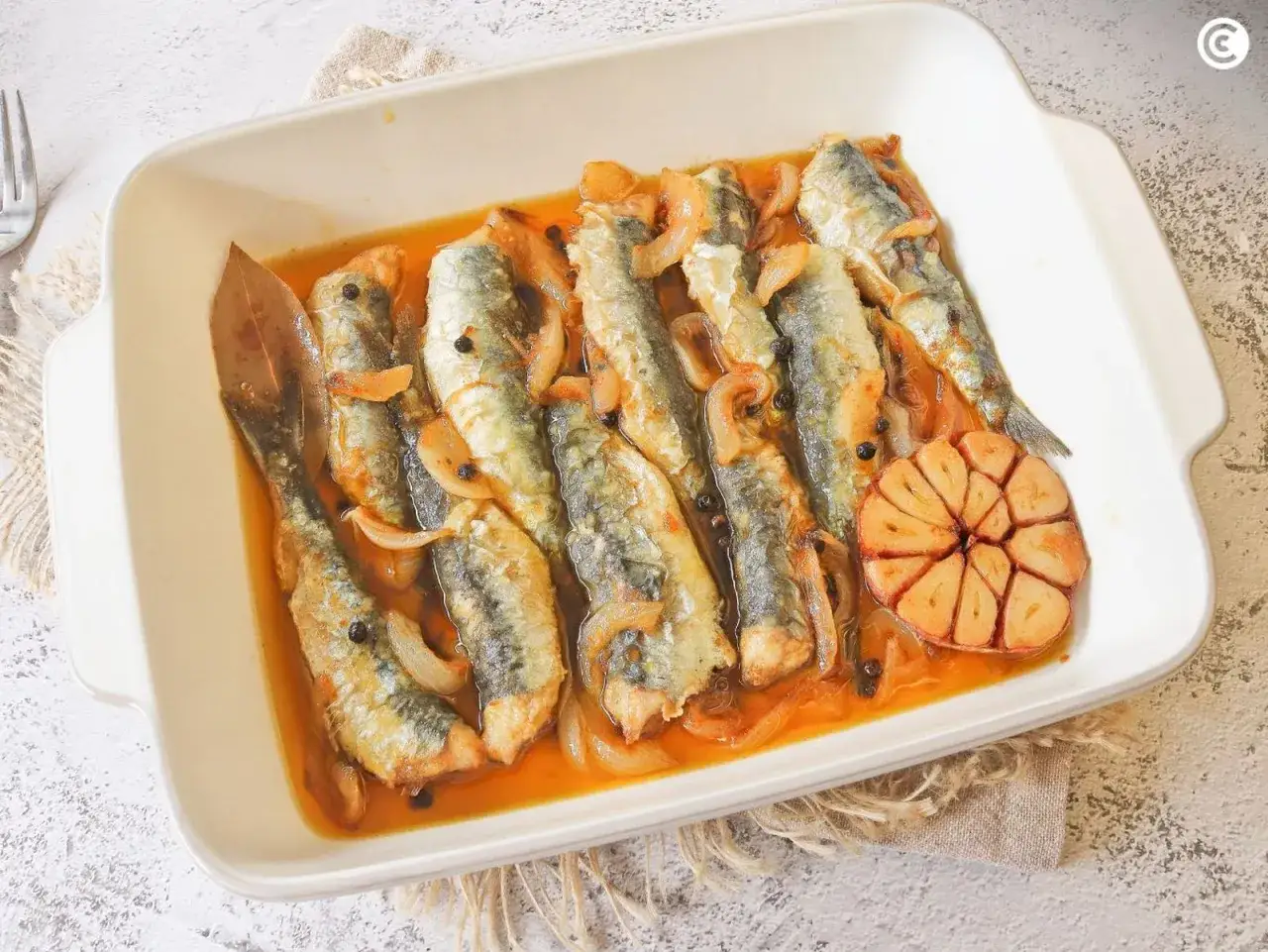 Sardinas en escabeche, con cebolla, granos de pimienta y un diente de ajo asado, servidas en una fuente blanca.